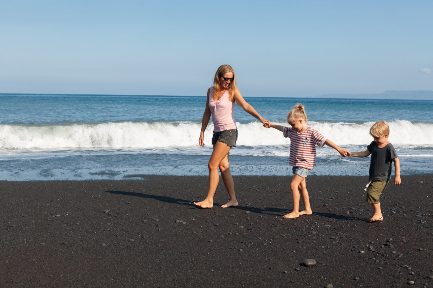 Happy family – young mother, daughter, baby son hold hands, walk together by water pool along sea surf on black sand beach. Travel, active lifestyle, parents with child on summer vacation