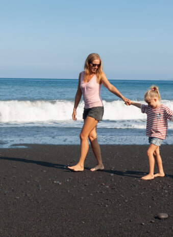 Happy family - young mother, daughter, baby son hold hands, walk together by water pool along sea surf on black sand beach. Travel, active lifestyle, parents with child on summer vacation