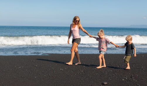 Happy family - young mother, daughter, baby son hold hands, walk together by water pool along sea surf on black sand beach. Travel, active lifestyle, parents with child on summer vacation